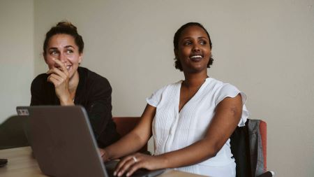 två kvinnor sitter bredvid varandra, vid ett bord med en dator