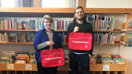 En manlig och en kvinnlig person står i ett bibliotek. De håller upp varsin röd väska med texten Logopedväska på.