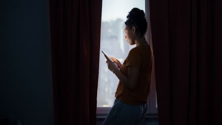 Kvinnlig person står i glipan mellan två gardiner och tittar på sin telefon.