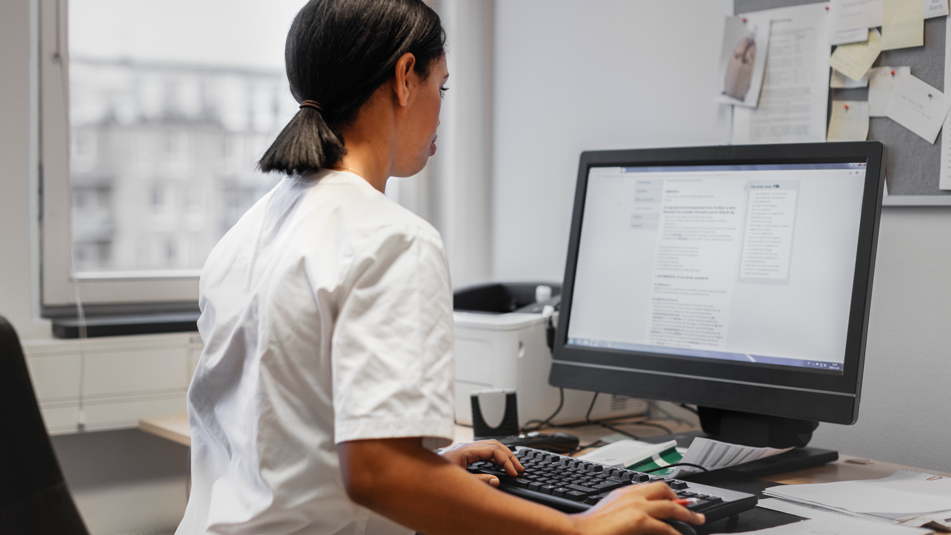 Kvinna i vita vårdkläder sitter framför en dator vid ett skrivbord