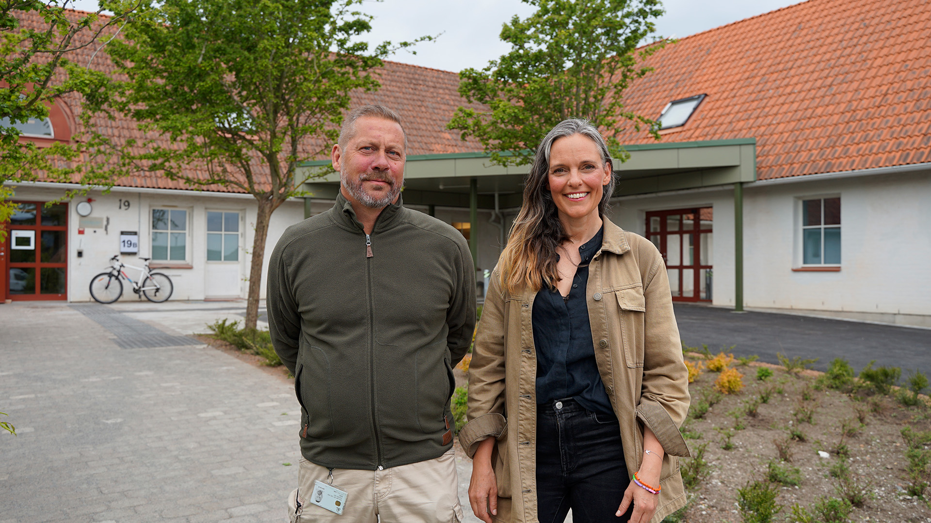 En och en kvinna står framför ett hus och tittar in i kameran. 