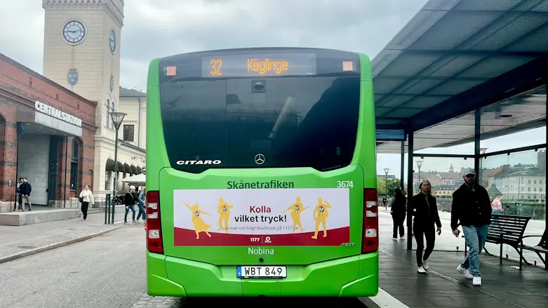 Baksidan av en grön stadsbuss vid Malmö Centralstation där det står Kolla vilket tryck på annonsplatsen på bussens baksida.