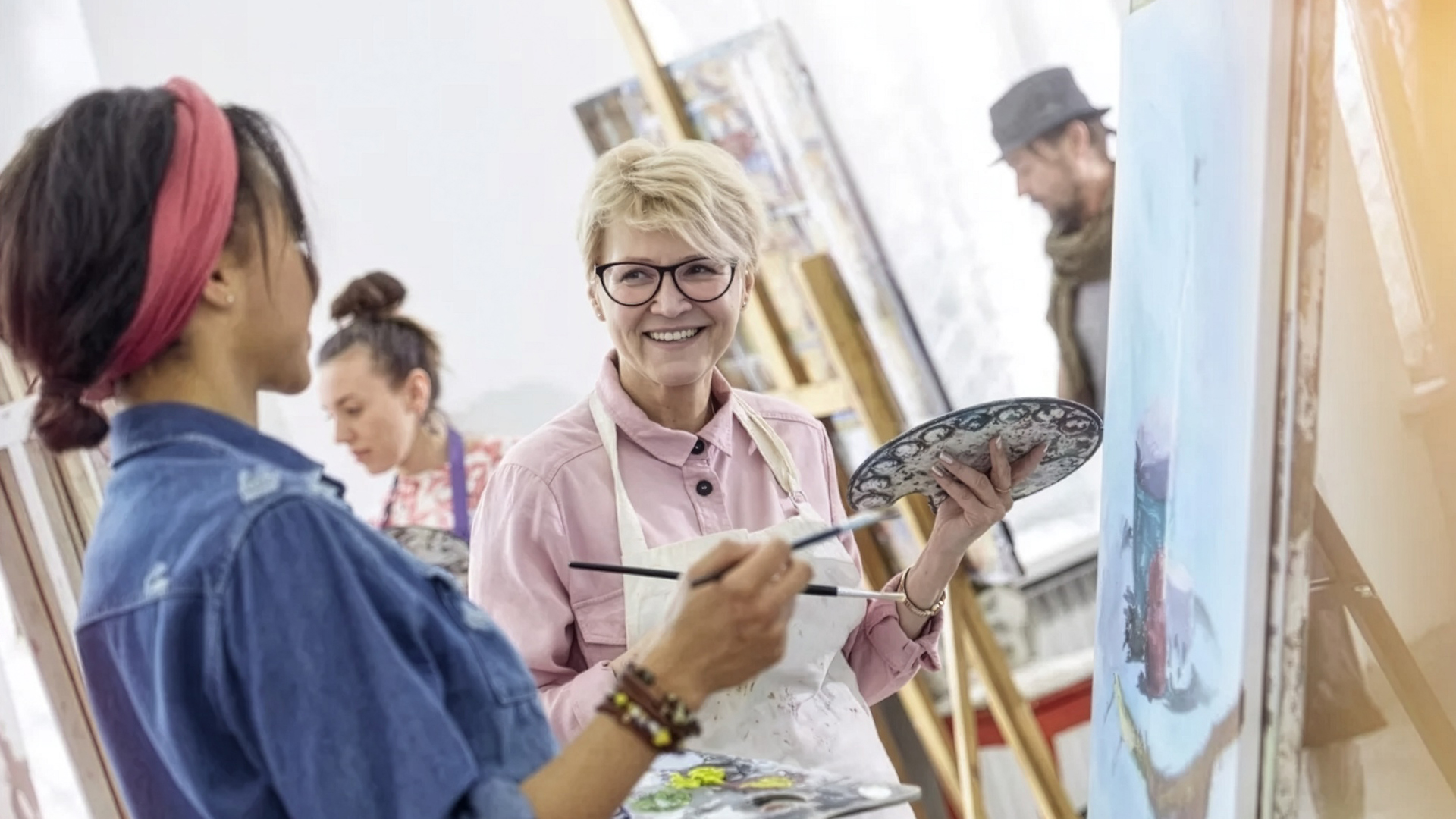 Två kvinnor står framför ett staffli med målarpenslar. I bakgrunden syns ytterligare två personer som målar