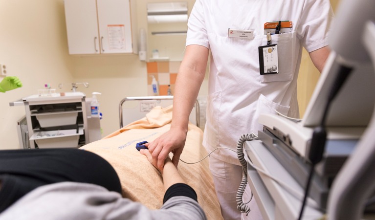 Vitklädd vårdpersonal tar pulsen på patient som ligger i säng.