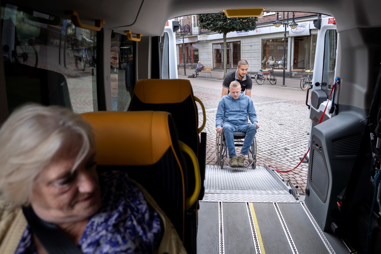 Inifrån en sjuktransport, en äldre kvinna sitter i bilen , en yngre man i rullstol rullas in bak i bilen. 