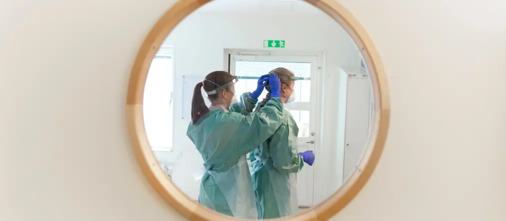 Två kvinnor hjälper varandra ta på skyddsutrustning i vården.