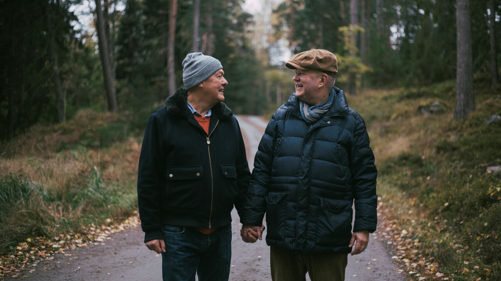 Ett äldre homosexuellt par håller varandra i  handen under en skogspromenad