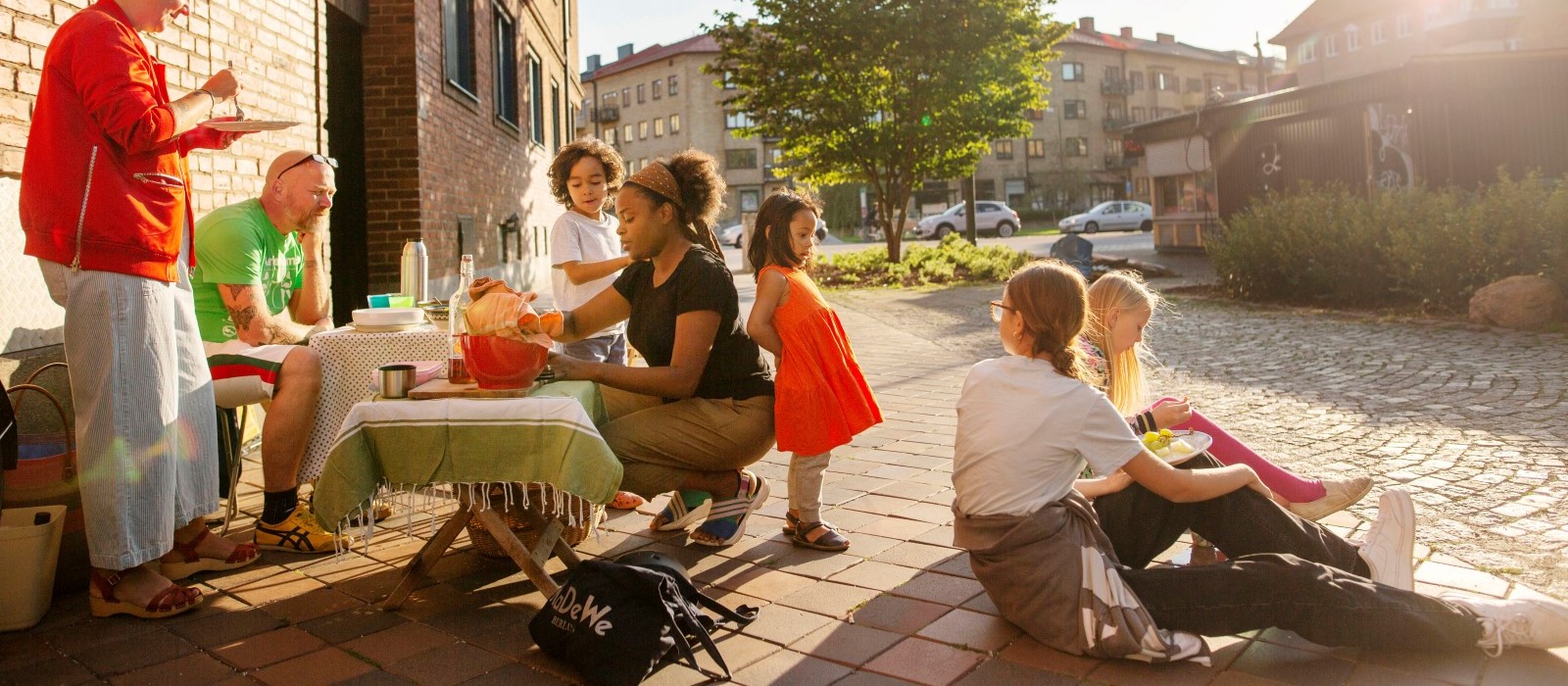 Grannar sitter på en trottoar och delar en måltid. 