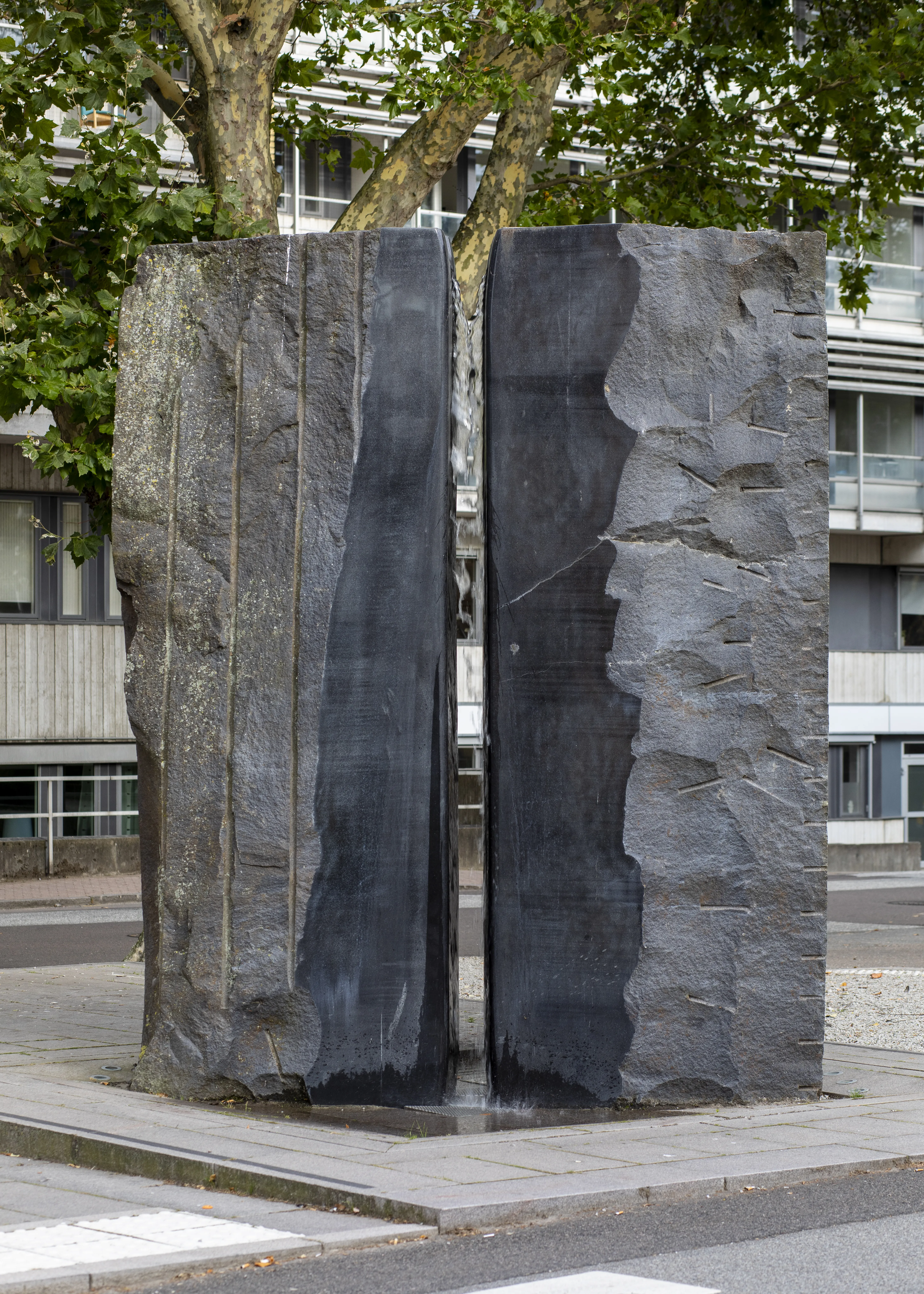 Två stora stenblock i svart granit står med ett litet mellanrum. Längs med stenarnas mitt rinner vatten ned som en fontän och fyller mellanrummet.