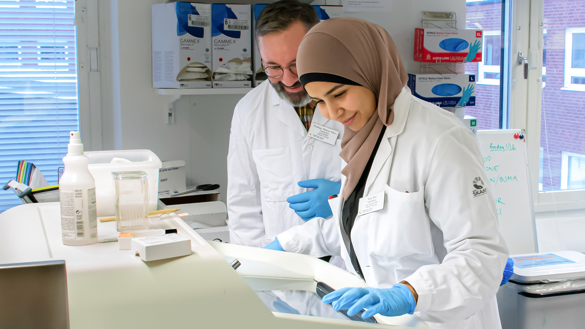 En manlig och en kvinnlig person  klädda i labbrockar arbetar i labbmiljö.