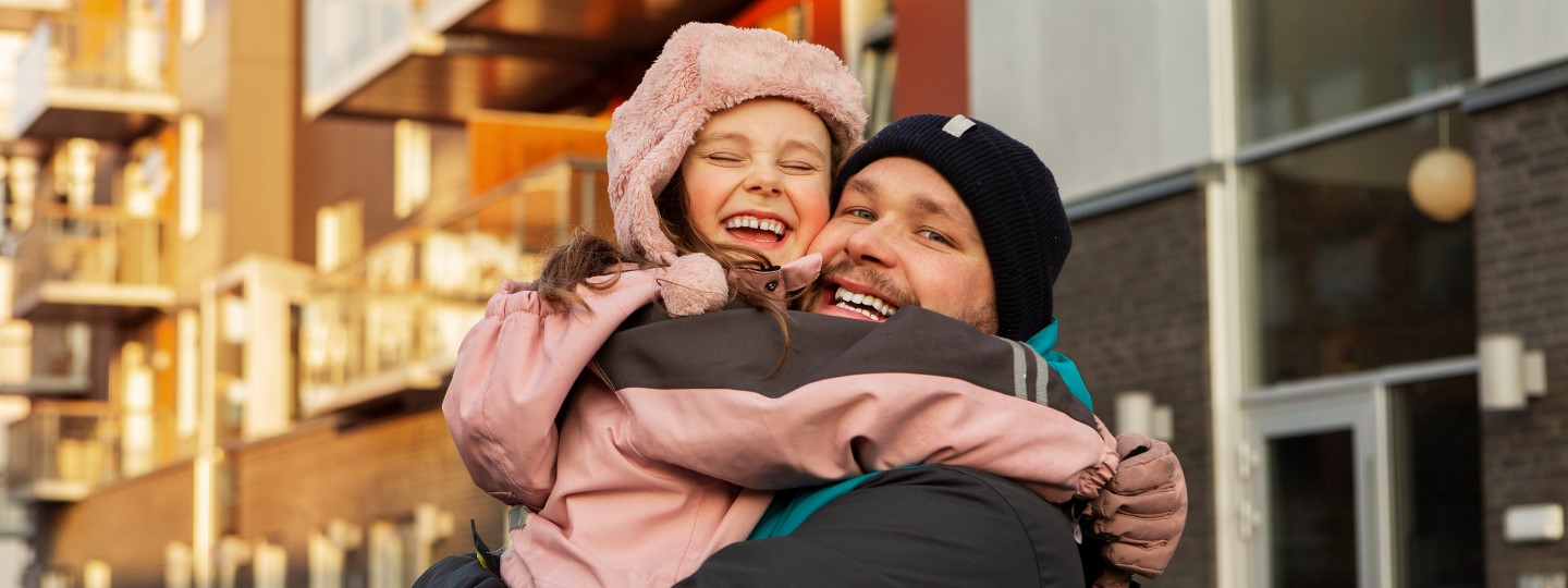 En pappa och ung dotter kramar om varandra utomhus och ser glada ut. De har på sig jackor och mössor.