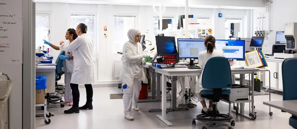 En sal med datorer och laboratorieutrustning. Fyra personer arbetar med olika saker. En sitter vid datorn, en håller i prover, och två diskuterar något.