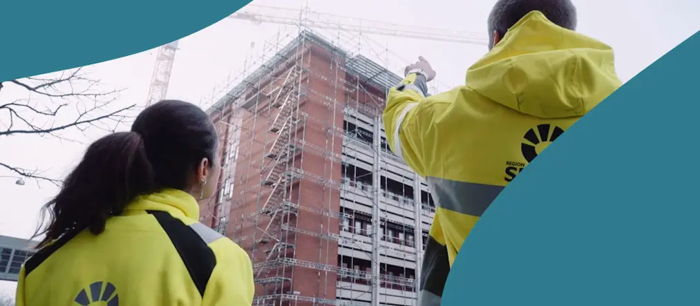 En kvinna och en man i varselkläder pekar och tittar mot ett husbygge