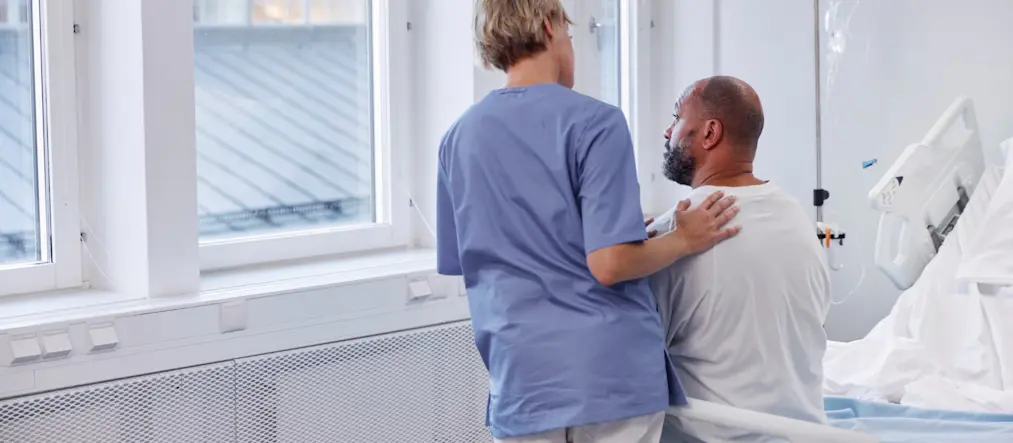 En sjuksköterska klappar en patient på ryggen, de tittar ut genom ett sjukhusfönster.