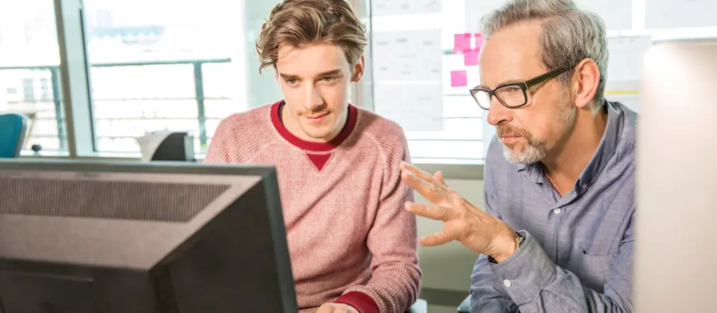 Två män sitter framför en dator. Den äldre mannen förklarar något för den yngre mannen.