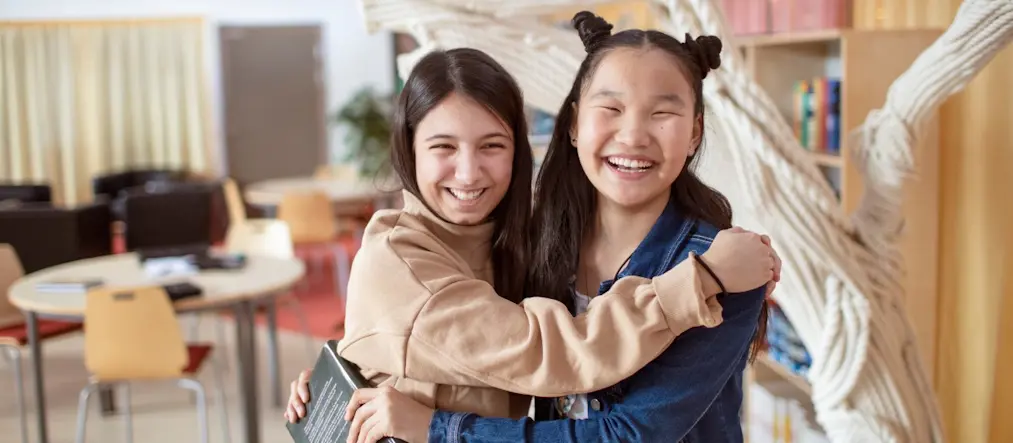 Två unga tonårstjejer tittar glatt in i kameran och håller om varandra. Bakgrunden är en skolmiljö.