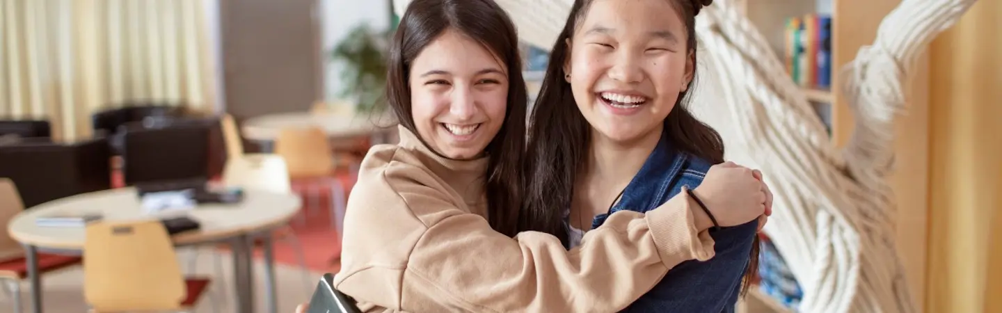 Två unga tonårstjejer tittar glatt in i kameran och håller om varandra. Bakgrunden är en skolmiljö.