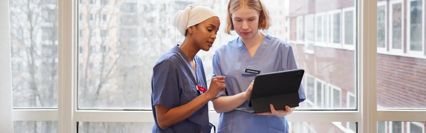 Två kvinnor i sjukhuskläder står inomhus framför ett stort fönster och tittar på en iPad.