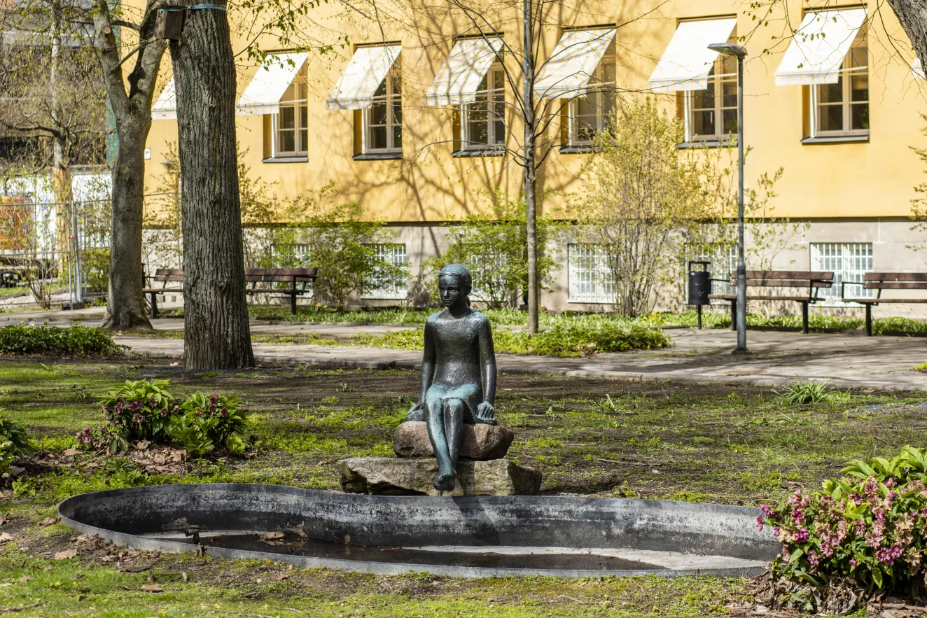 En flicka sitter på en sten, hennes ben hänger ner mot vattnet i en liten damm. Skulpturen av flickan är gjord i brons som ärgat och nu ser grönblå ut. 