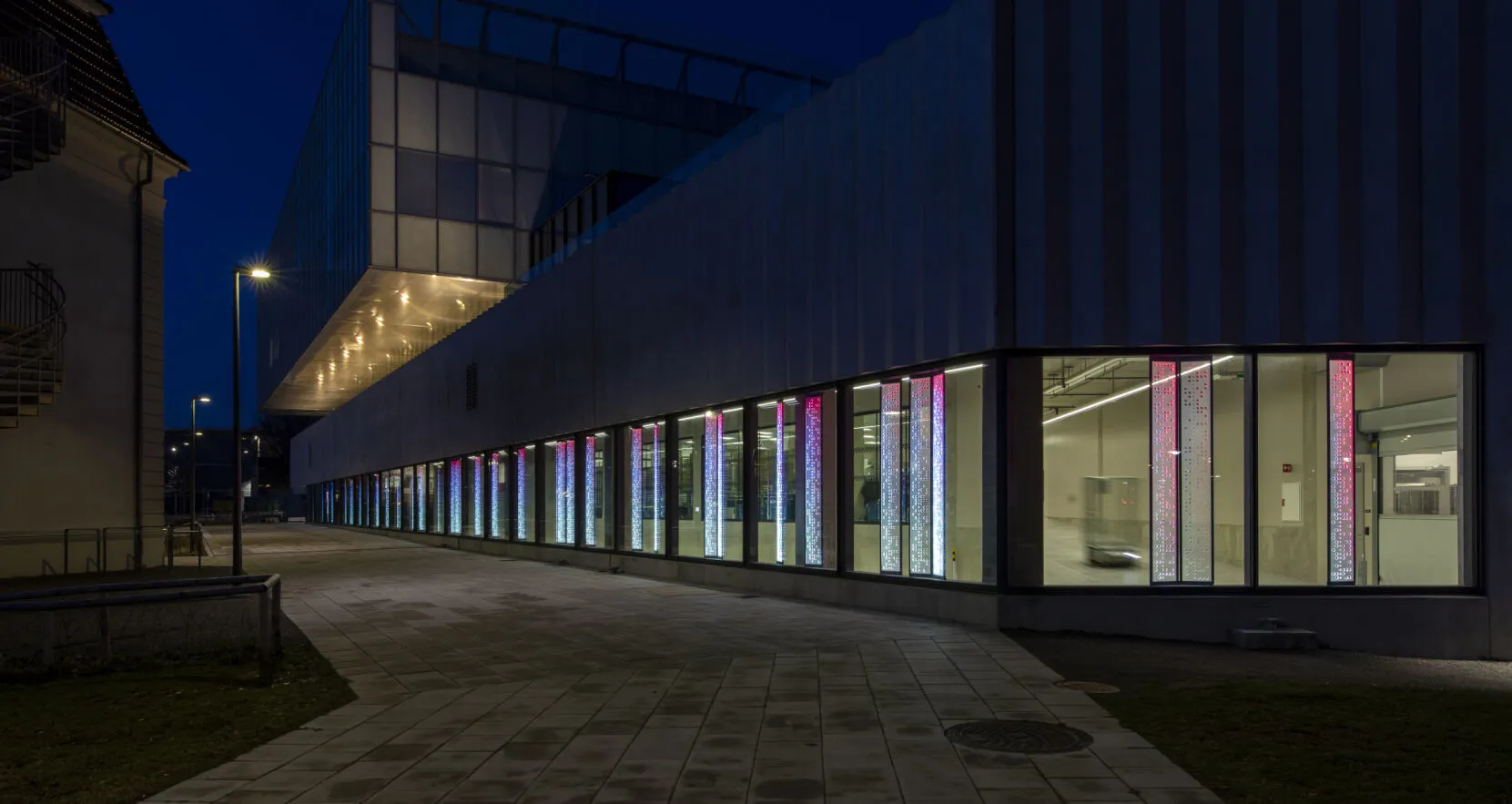 Det är kväll och det lyser inifrån fönsterna på servicebyggnaden i Malmö. I varje fönster syns en ljusinstallation där ljuset faller från lila färg i toppen till blått i botten. 