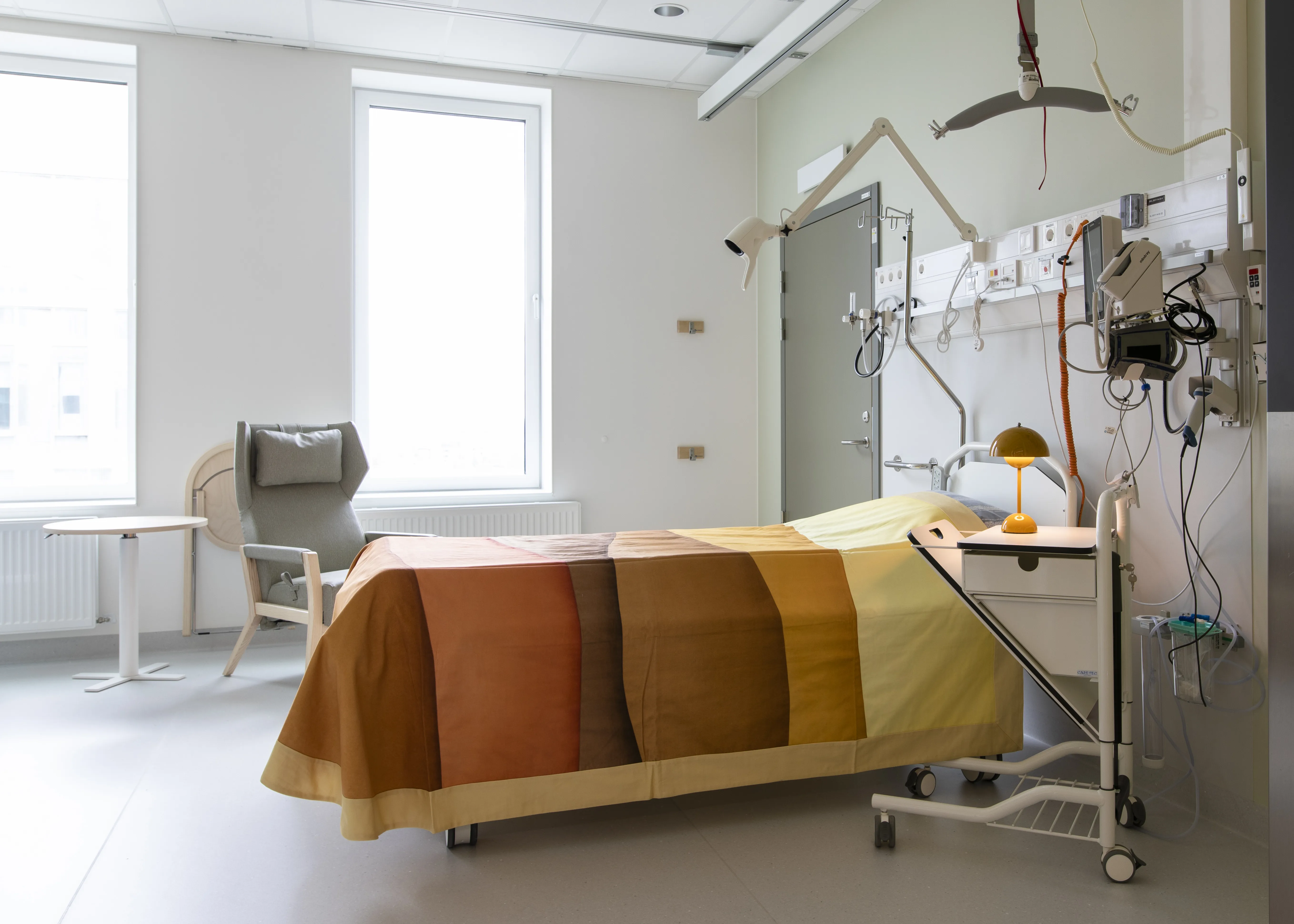 Inne i ett vårdrum står en bäddad säng, bredvid sängen står ett bord där en lampa är tänd. Sängen är bäddad med ett täcke vars färger påminner om en solnedgång, från ljust gul till bränd orange. 
