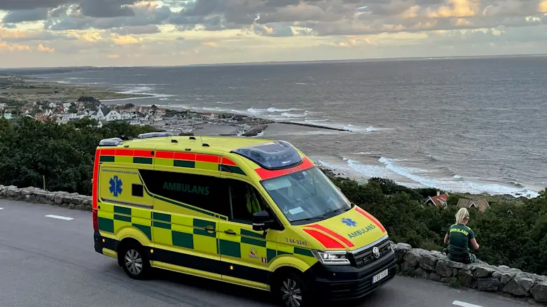 en ambulans är parkerad vid sidan av en väg som har utsikt över havet. En kvinnlig ambulanssjukvårdare sitter på en mur bredvid och tittar ut över havet.