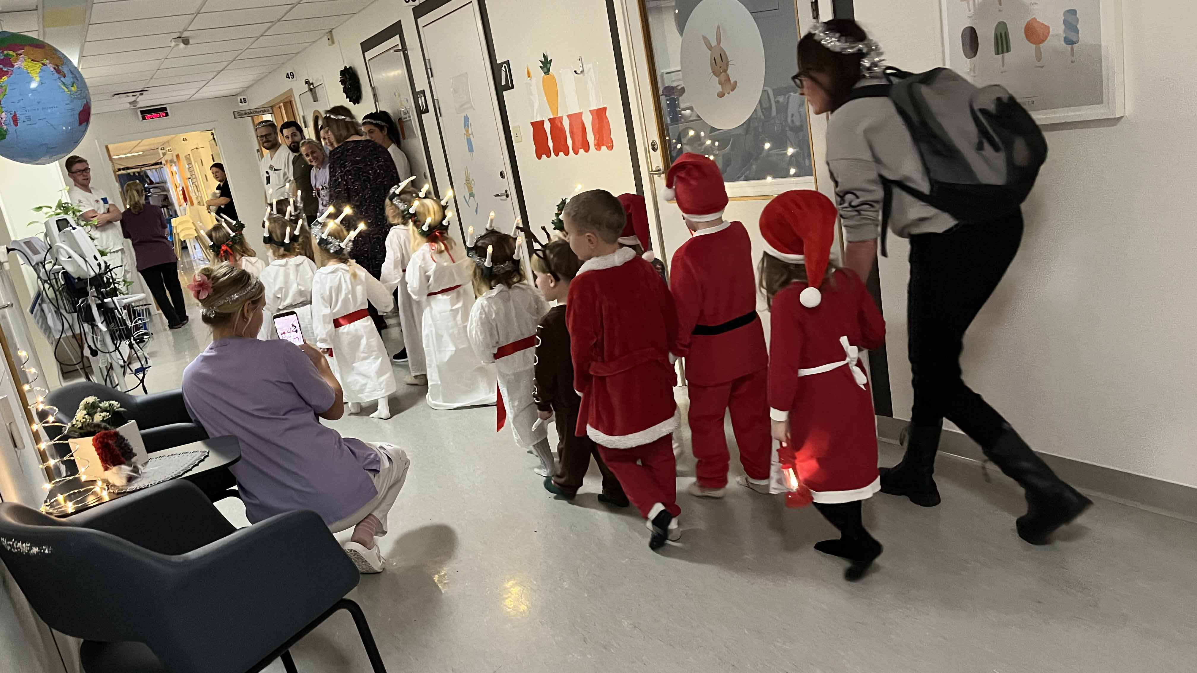 förskolebarn klädda i lucia- och tomtedräkter går luciatåg i en sjukhuskorridor. Medarbetare står längs väggarna.