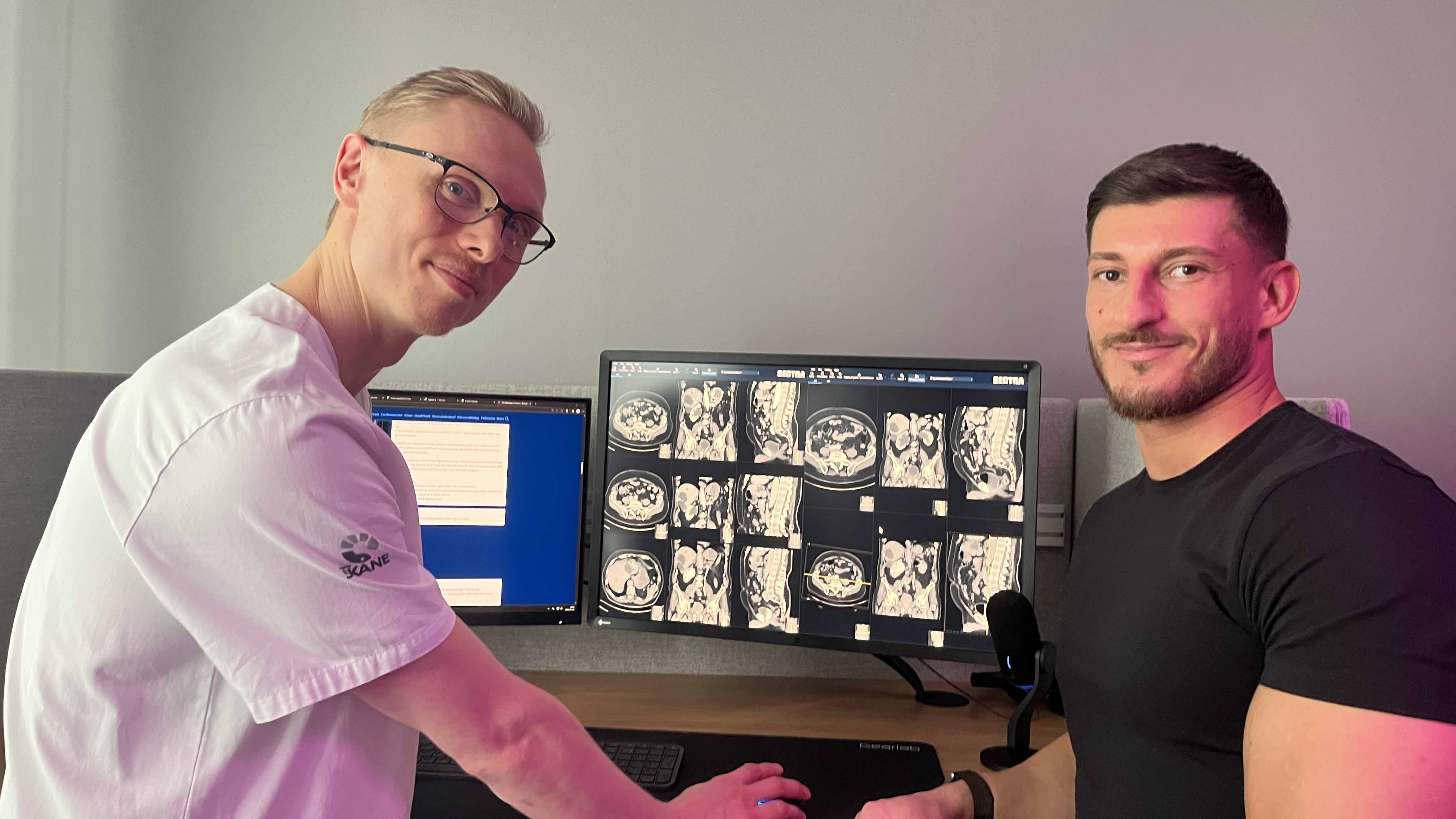 Oskar ERiksson, ST-läkare, i vita vårdkläder och Ismar Zejnic, beskrivande radiograf, i svart t-shirt, står vid ett skrivbord med en bildskärm med röntgenbilder.