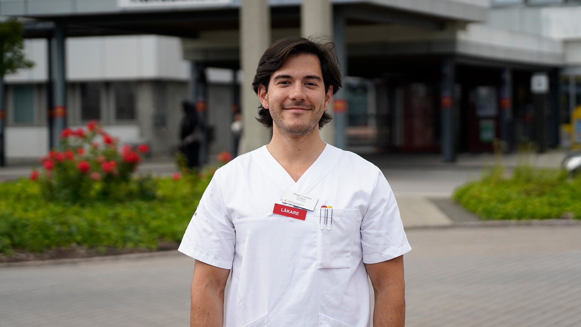 Läkaren Diego Cardeña Rodriguez står utanför Centralsjukhuset Kristianstad klädd i vita vårdkläder, tittar in i kameran.