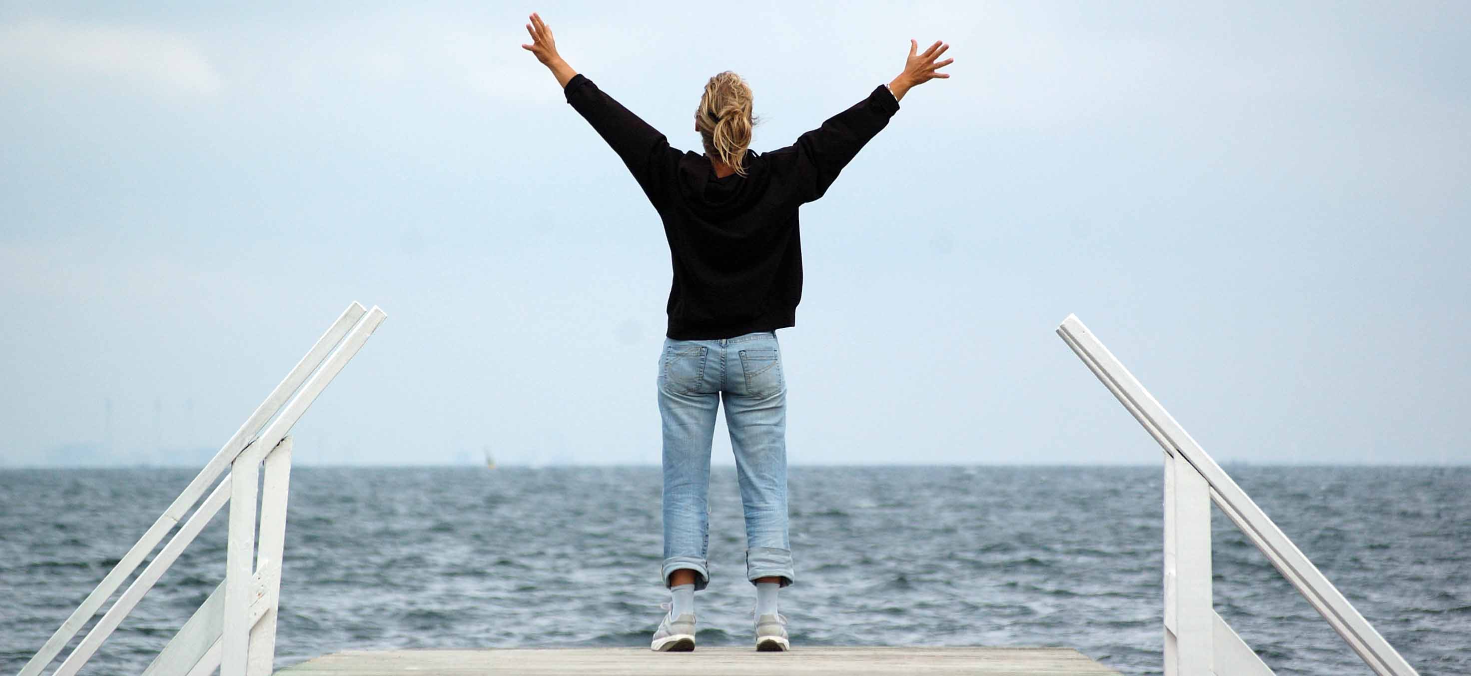 Kvinna fotograferad bakifrån står längst ut på en brygga med armarna i luften