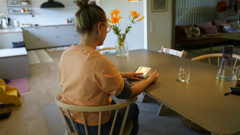En kvinna sitter med ryggen mot kameran vid ett matbord och mäter sitt blodtryck med hjälp av en digital mätare.
