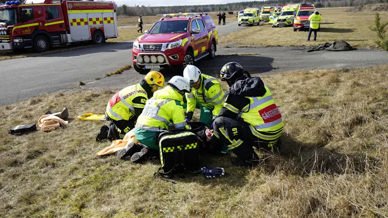 en grupp räddningspersonal klädda i gula reflexvästar och hjälmar sitter på knä runt om en person som spelar skadad i en övning. I bakgrunden ett fält med flera parkerade ambulanser och andra räddningsfordon.