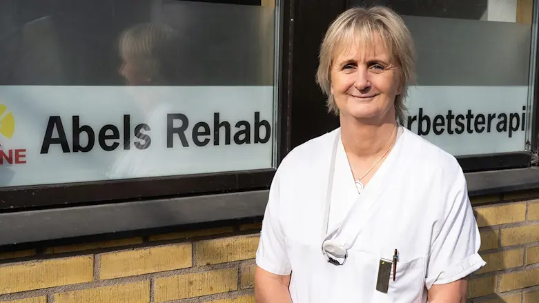Porträttfoto av fysioterapeut Malin Jönsson Lundgren iklädd vita arbetskläder framför en brun tegelvägg med texten Abels rehab i fönstret.