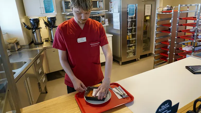 Servicevärden Felix Eriksson iklädd röd arbetsskjorta, står i ett storkök och lägger upp mat på en tallrik.
