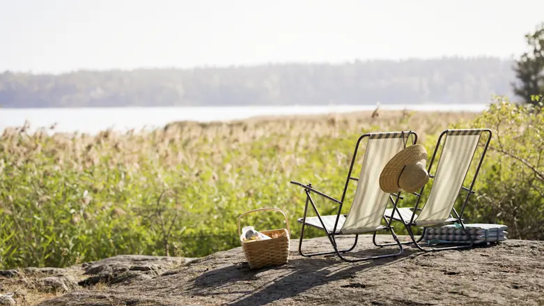 Fotografi av två solstolar som står uppställda på en klippa intill havet. Framför vattnet syns vass och intill solstolarna står en picknickkorg.