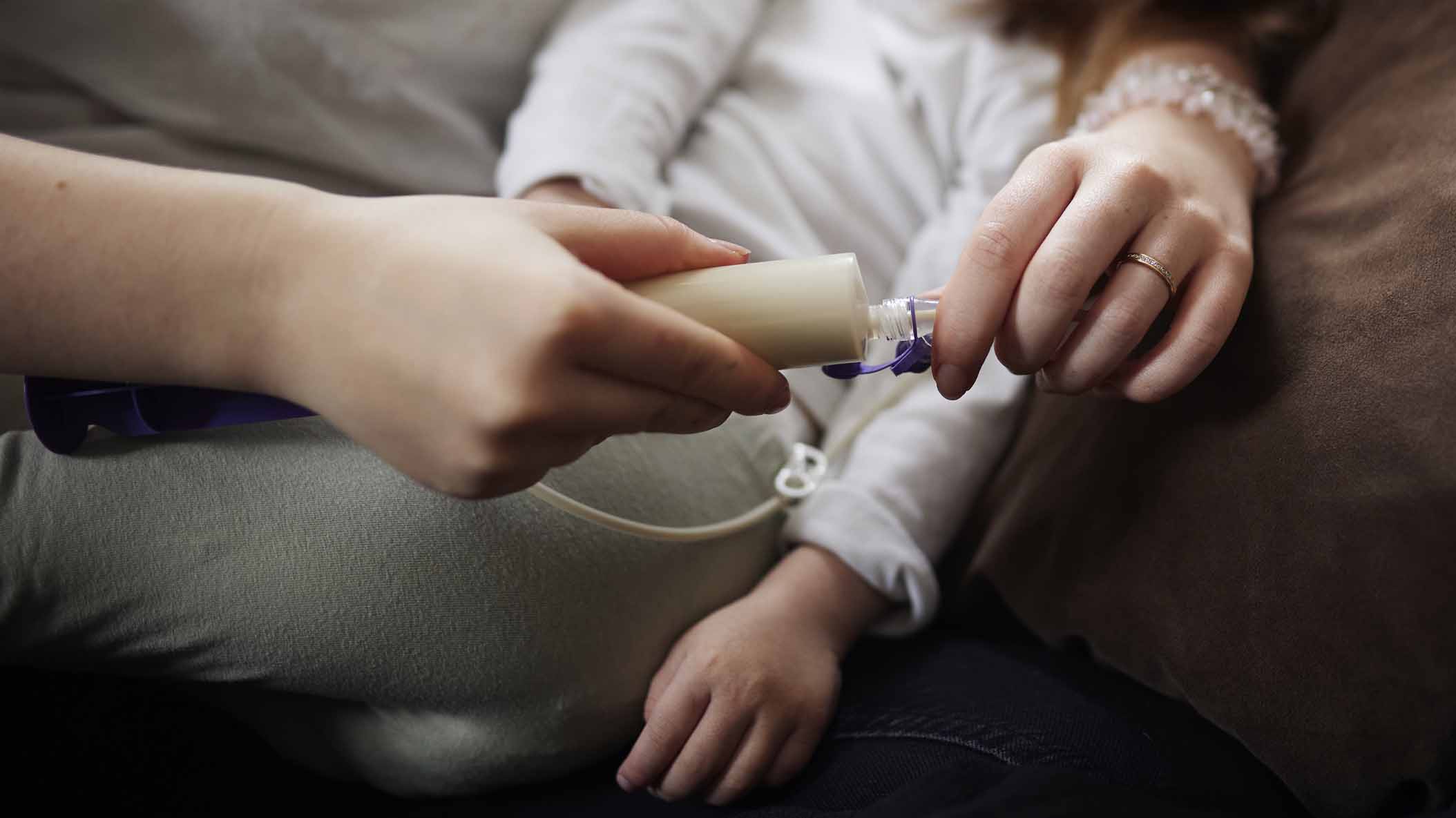 närbild på kvinna med litet barn som ligger i famnen. Mammans händer håller i en spruta för sondmat.