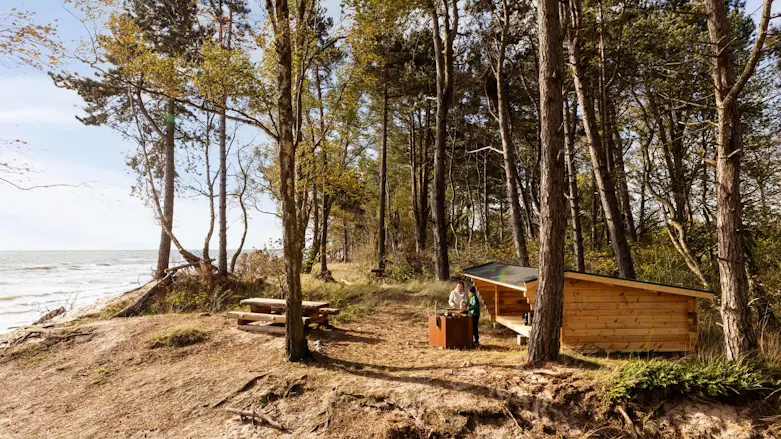 Foto av en bit kustremsa med tallskog, hav, ett vindskydd och två personer som står och grillar.