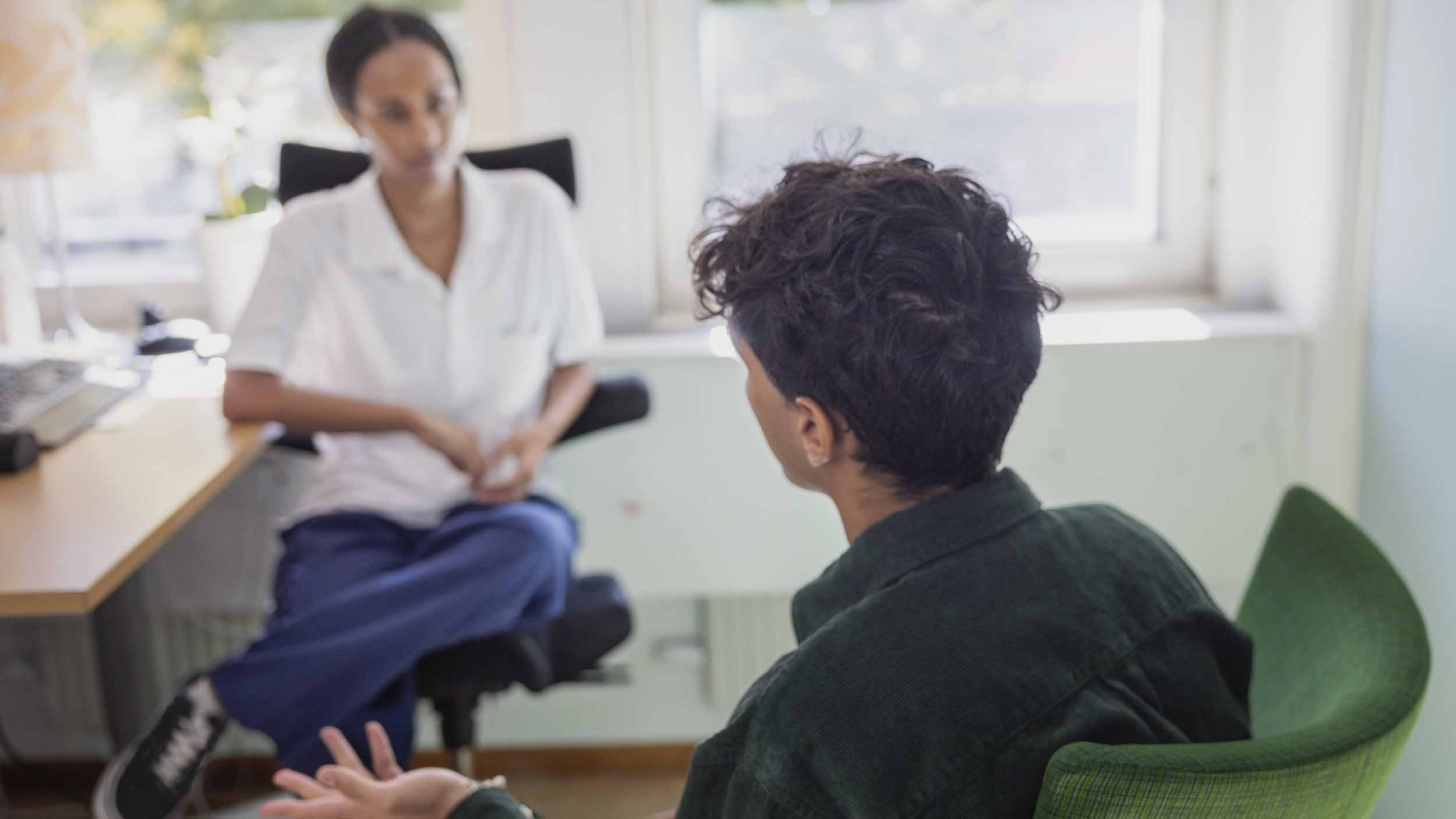 en ung man sitter mittemot en kvinnlig vårdmedarbetare i vita vårdkläder. Mannen är fotad snett bakifrån.