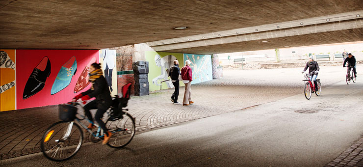 Tre cyklister och två personer förflyttar sig i en passage, i stadsmiljö.