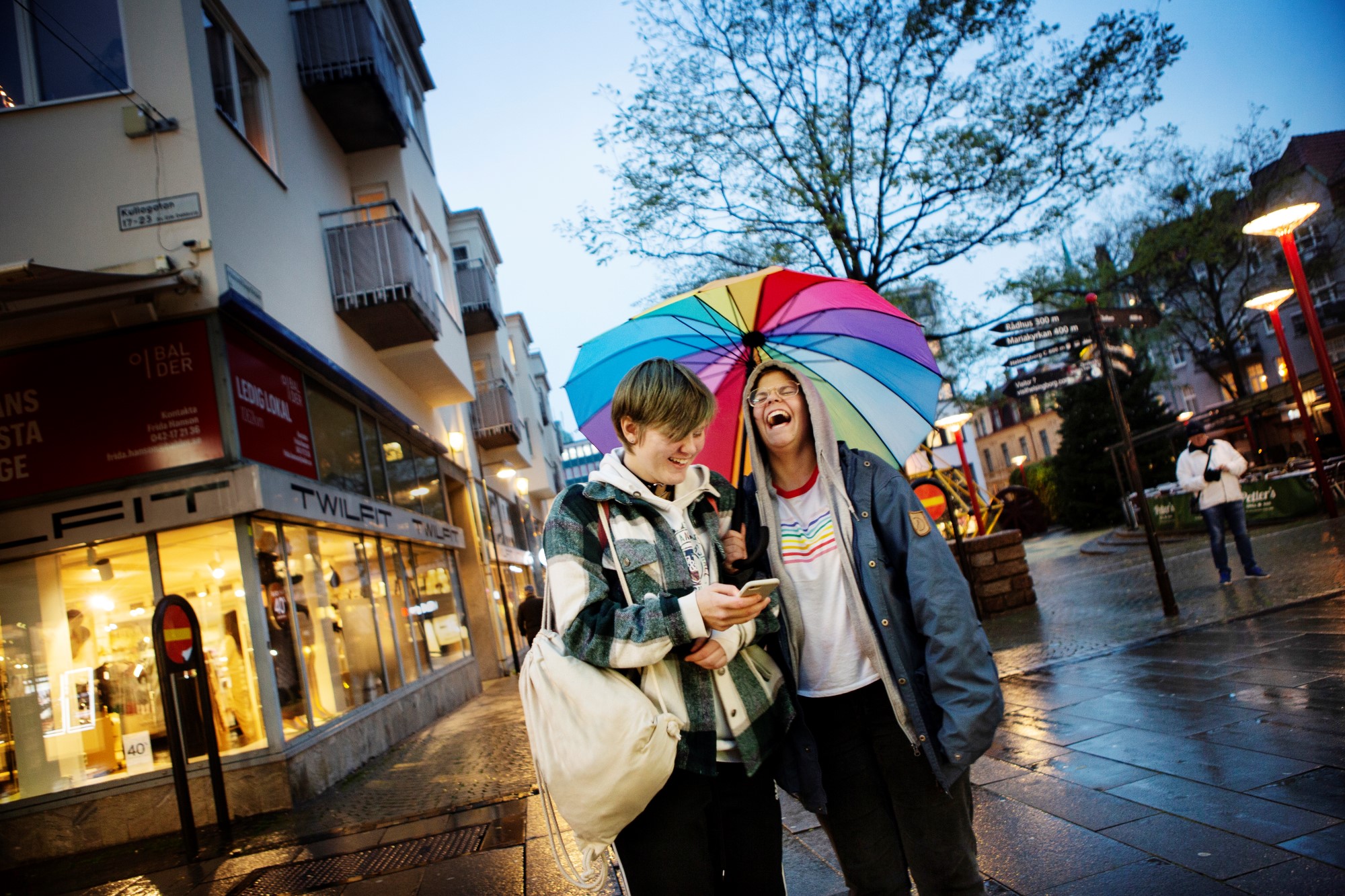 Två ungdomar ser glada ut under ett färgglatt paraply i stadsmiljö.